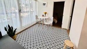 Ein kleiner, gut beleuchteter Raum mit einem gemusterten Boden, einem runden Tisch mit Stühlen, einem Blumentopf und einer Tür zu einem anderen Raum.