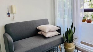 Ein graues Sofa mit weißen Kissen, ein Blumentopf und ein gemustertes Teppich in einem Zimmer mit weißer Wand und einem Fenster mit durchsichtigen Vorhängen.