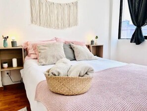 Une chambre bien rangée, avec un lit, des tables de chevet, une décoration murale en macramé et une fenêtre avec des rideaux.