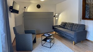 Een moderne woonkamer met een grijze bank, een kleinere fauteuil, een salontafel en een aan de muur bevestigde televisie.