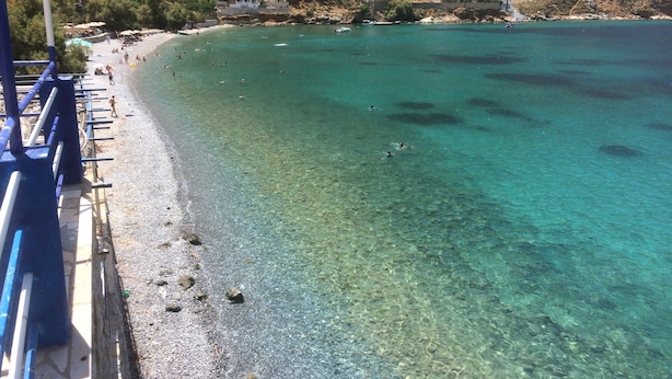 Ein Küstenstrand mit klarem türkisblauem Wasser, felsigem Gelände und verstreuten Gebäuden.