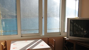 Ein Zimmer mit einem Tisch, bedeckt mit einer weißen Tischdecke mit orangefarbenen Mustern, zwei Holzstühlen, einem Fernseher auf einem Holzschrank und einem Fenster mit Bergblick.