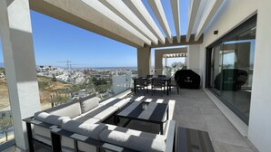 Un balcon aménagé avec un canapé, une table et un barbecue, offrant une vue sur le paysage urbain et la mer.