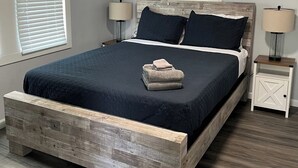 A bedroom with a wooden bed frame, a dark blue bedspread, two bedside lamps, and a small white nightstand with a cross-cut design.