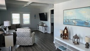 A modern living room with a sofa, a patterned ottoman, a TV, a painting of sailboats, and a white console table with decorative items.