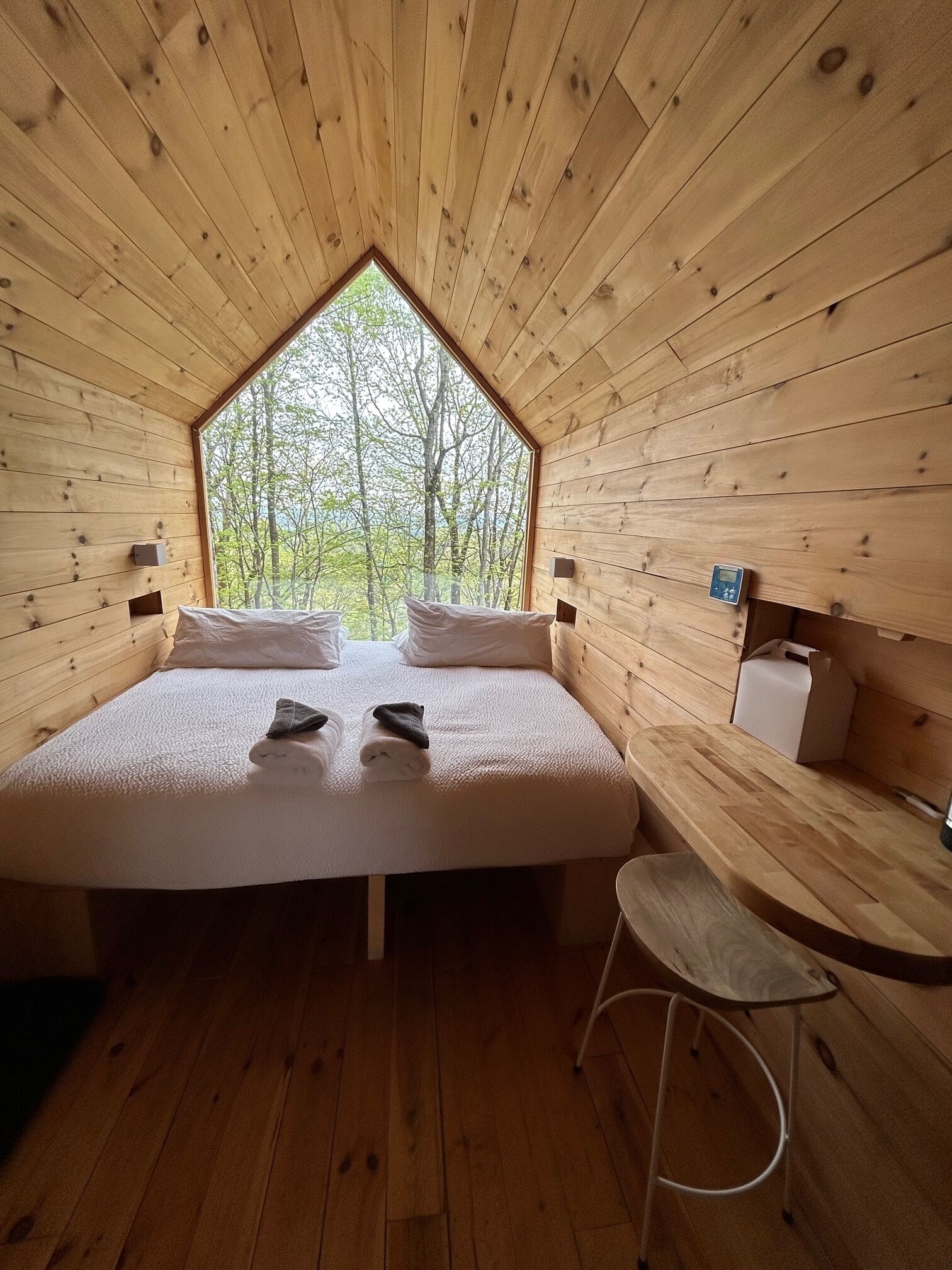 A wooden cabin bedroom with a large bed, a small table, and a view of trees through a window.