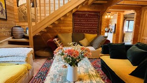 A cozy cabin interior with a wooden staircase, a sofa, a table with a vase of flowers, and a patterned rug.