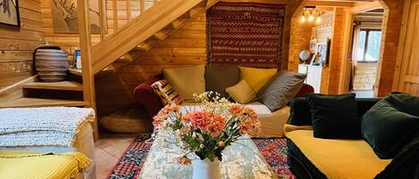 A cozy cabin interior with a wooden staircase, a sofa, a table with a vase of flowers, and a patterned rug.