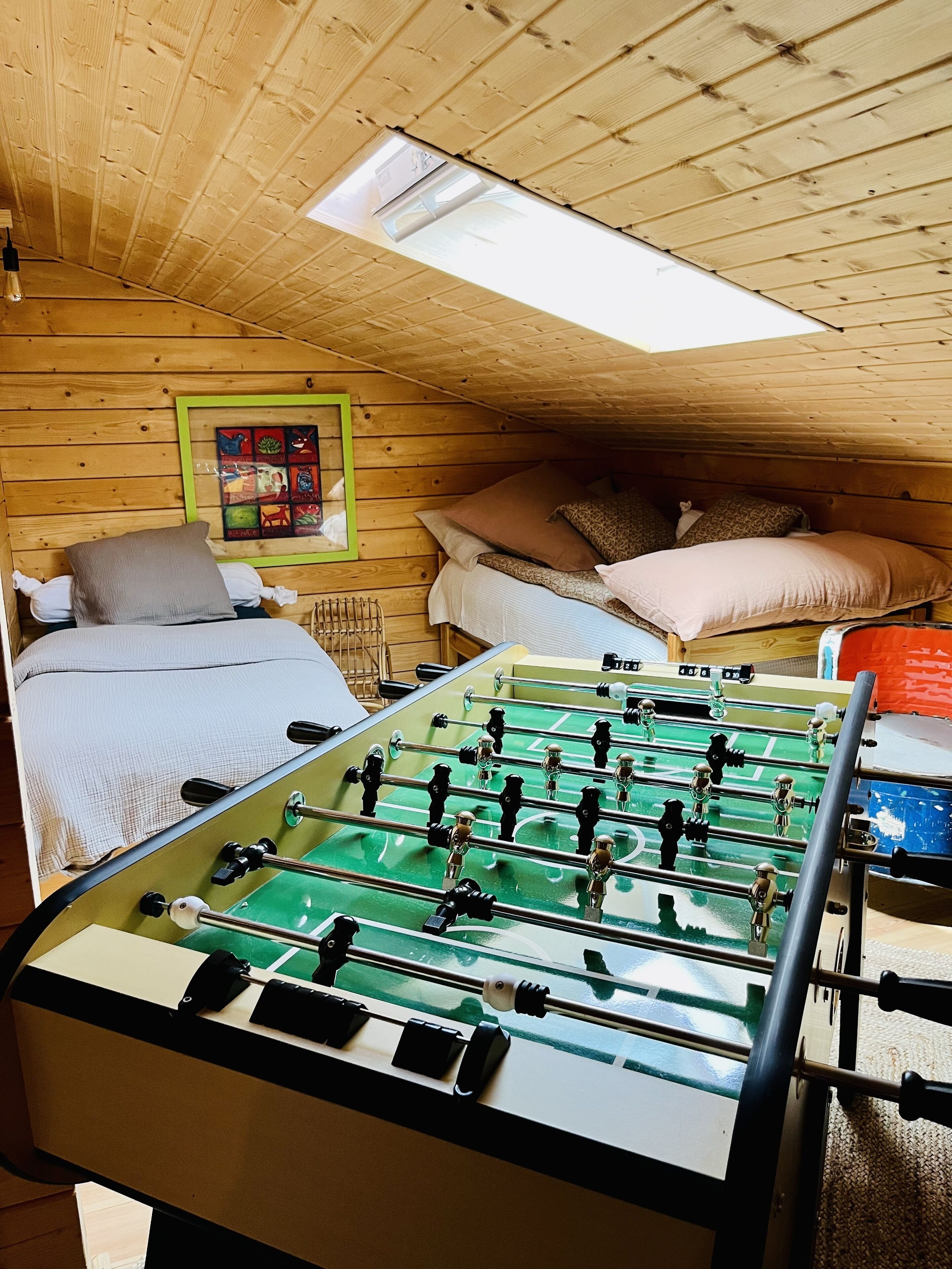 Ein gemütliches Zimmer mit Holzbalkendecke, einem Bett mit Kissen, einem Kicker und einem gerahmten Bild an der Wand.