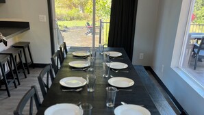 Een lange eettafel gedekt voor zes personen, met stoelen, een raam met uitzicht op een tuin en een donkerkleurige tafelkleed.