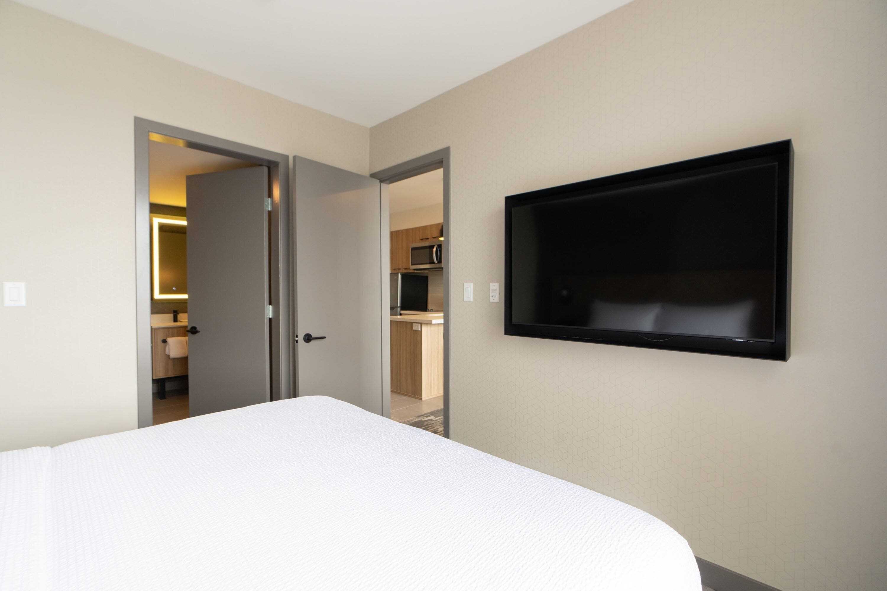 A hotel room with a bed, a television mounted on the wall, and an open door leading to a bathroom and kitchen area.