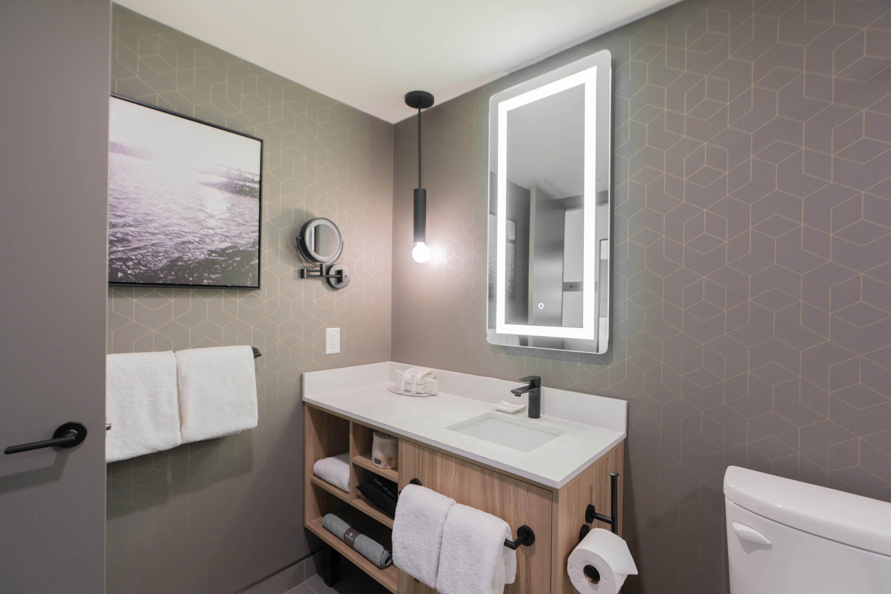 Een moderne badkamer met een witte wastafel, een spiegel en een toilet. Handdoeken hangen aan de muur en er is een plank met extra handdoeken.