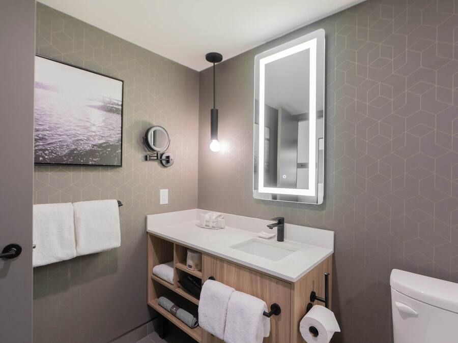 Een moderne badkamer met een witte wastafel, een spiegel en een toilet. Handdoeken hangen aan de muur en er is een plank met extra handdoeken.