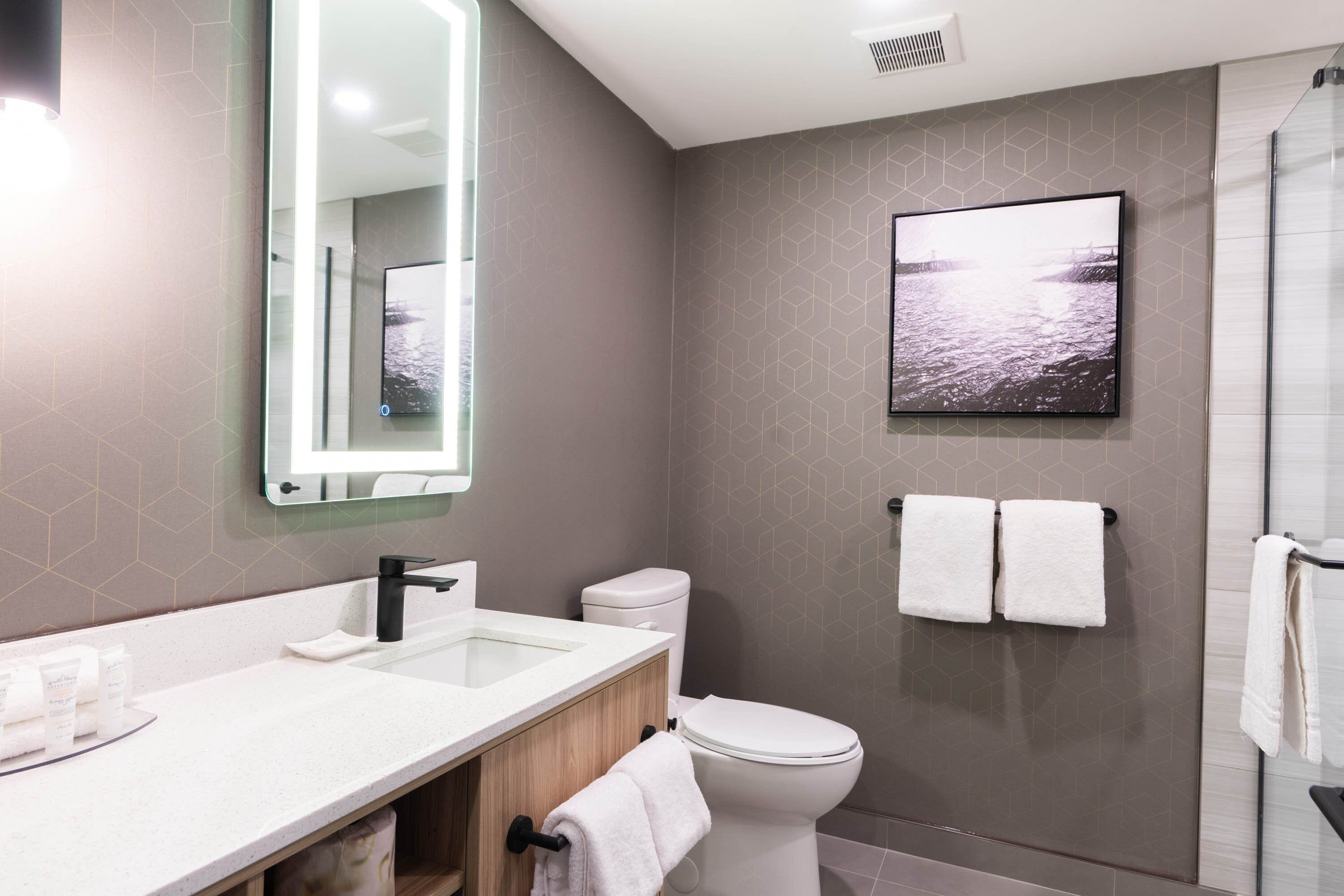 A modern bathroom with a large mirror, white sink, and toilet. There is a framed picture on the wall and two towels hanging on hooks.