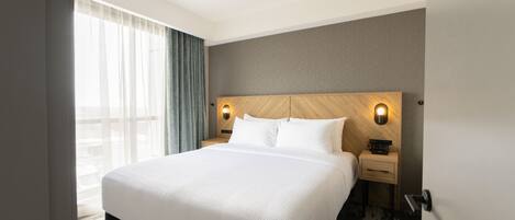 A hotel room with a large bed, a wooden headboard, a bedside table with a lamp, and a view of the outdoors through sheer curtains.