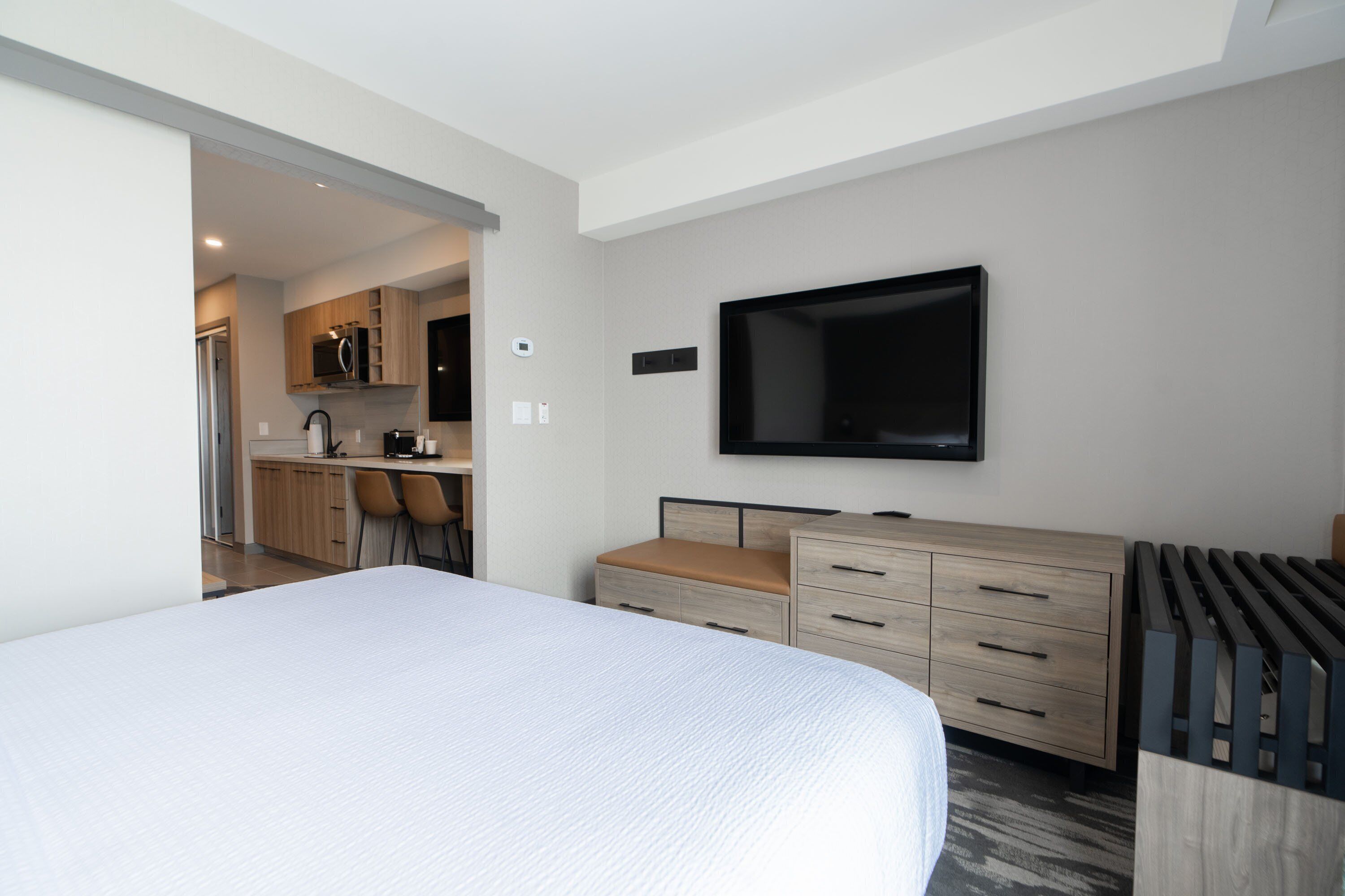 Een moderne hotelkamer met een groot bed, een aan de muur bevestigde televisie, een houten dressoir en een keukenruimte zichtbaar door een open deur.