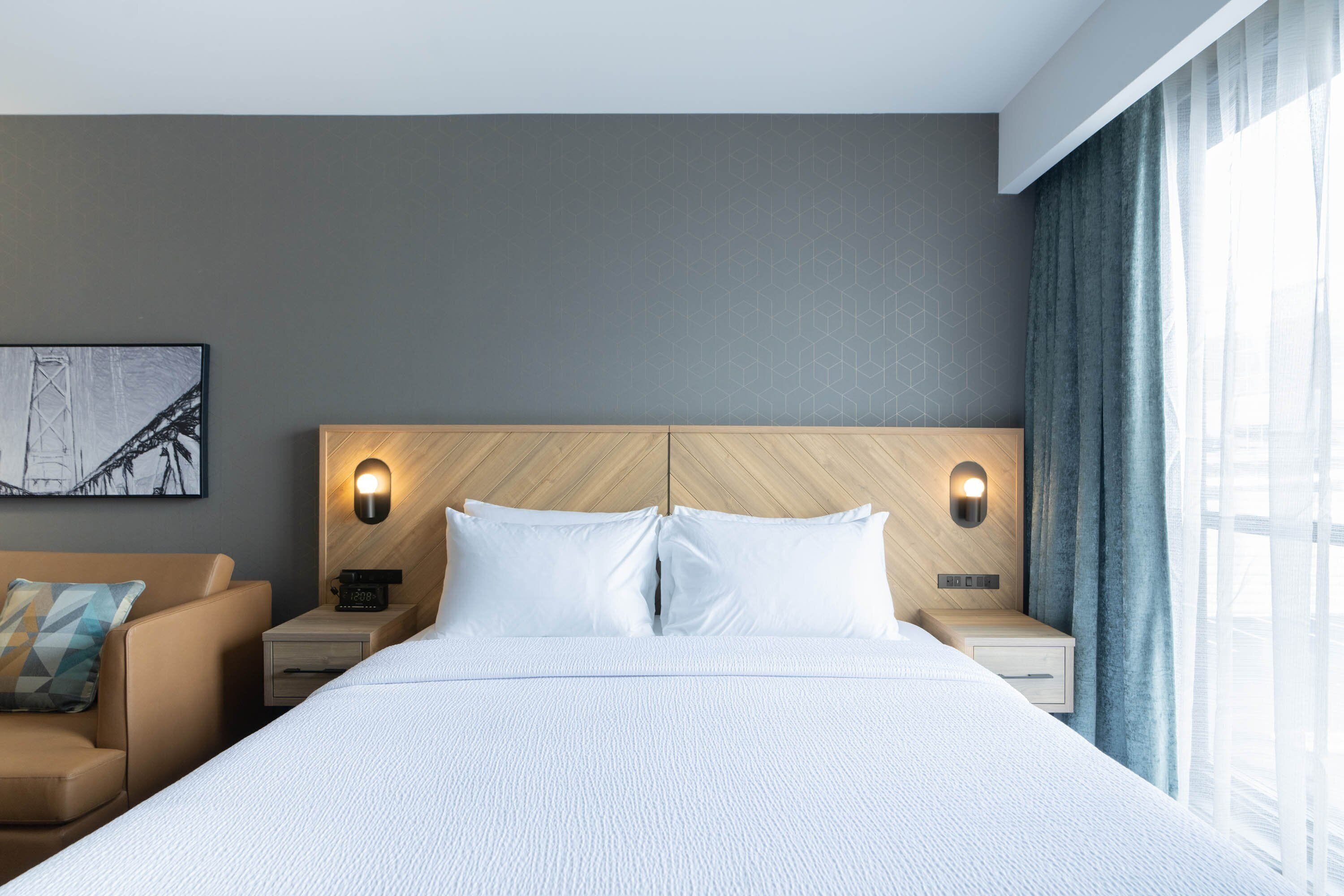 A modern hotel room with a large bed, two bedside tables with lamps, a headboard with geometric patterns, a framed picture on the wall, and a window with curtains.