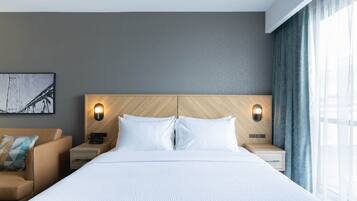 A modern hotel room with a large bed, two bedside tables with lamps, a headboard with geometric patterns, a framed picture on the wall, and a window with curtains.