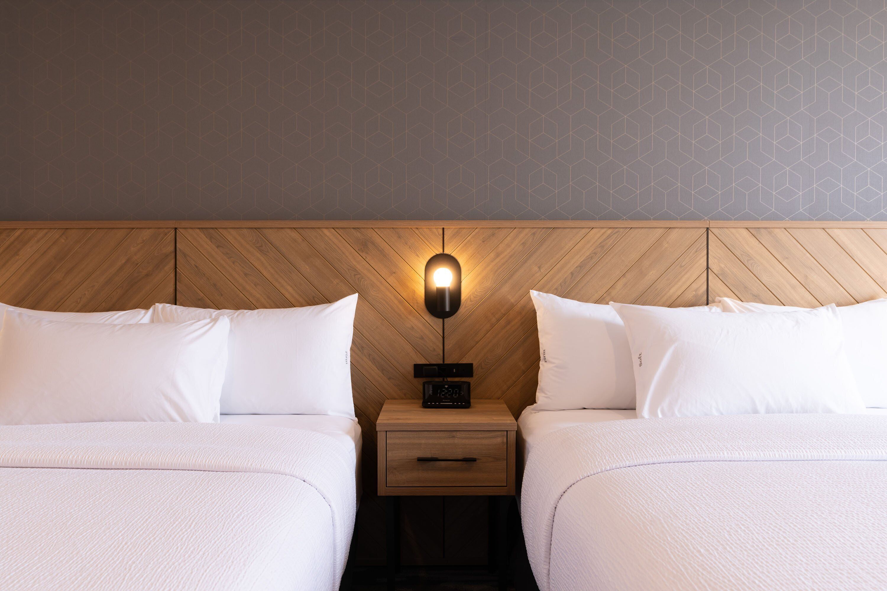 Habitación de hotel con dos camas, cabecero de madera, mesita de noche con un reloj y pared con diseño geométrico.