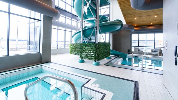 Una piscina interior con tobogán de agua, una sala iluminada por el sol con ventanas amplias y una pequeña zona de estar.