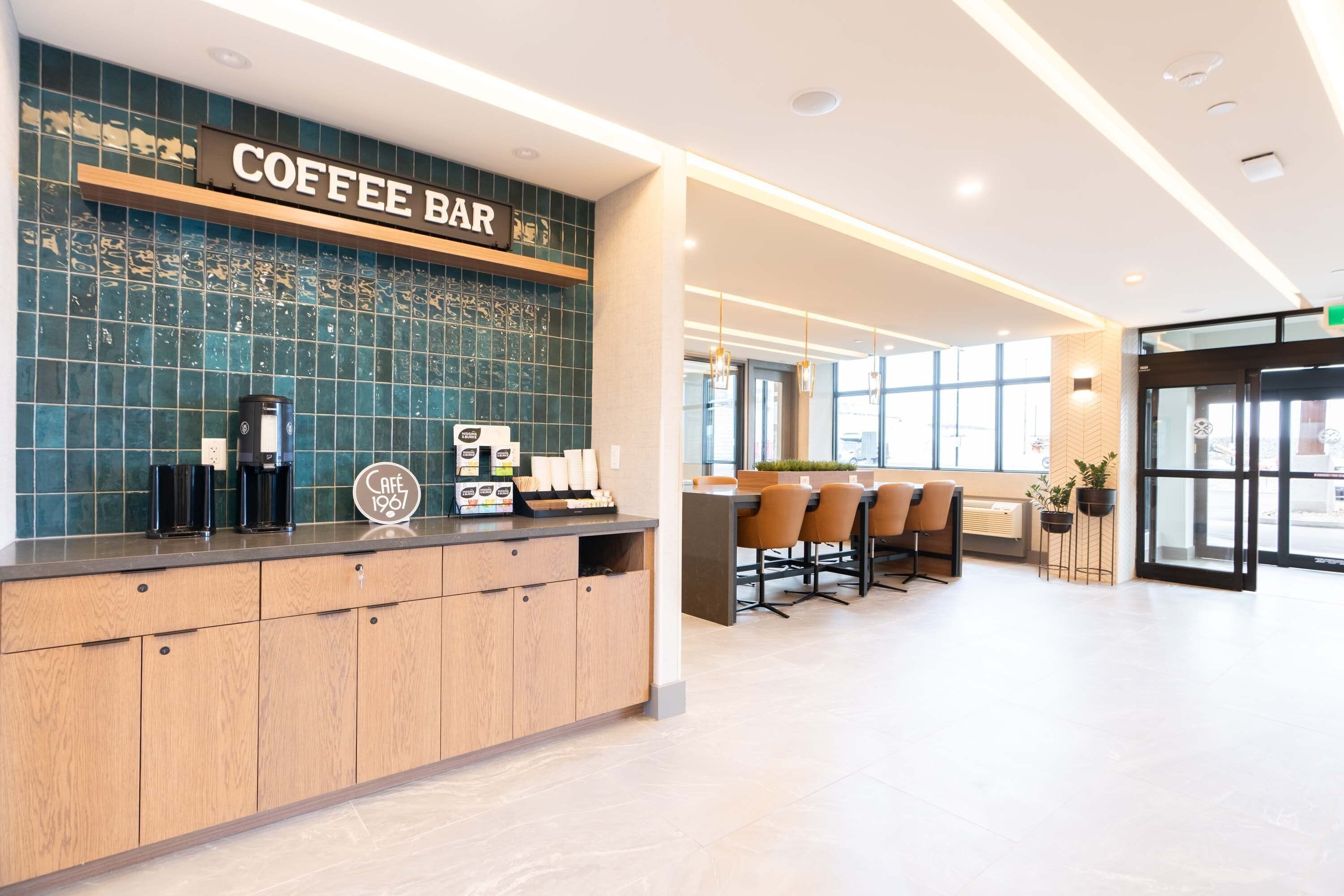 A coffee bar with a sign, espresso machines, and a menu display. There is a seating area with chairs and tables in the background.