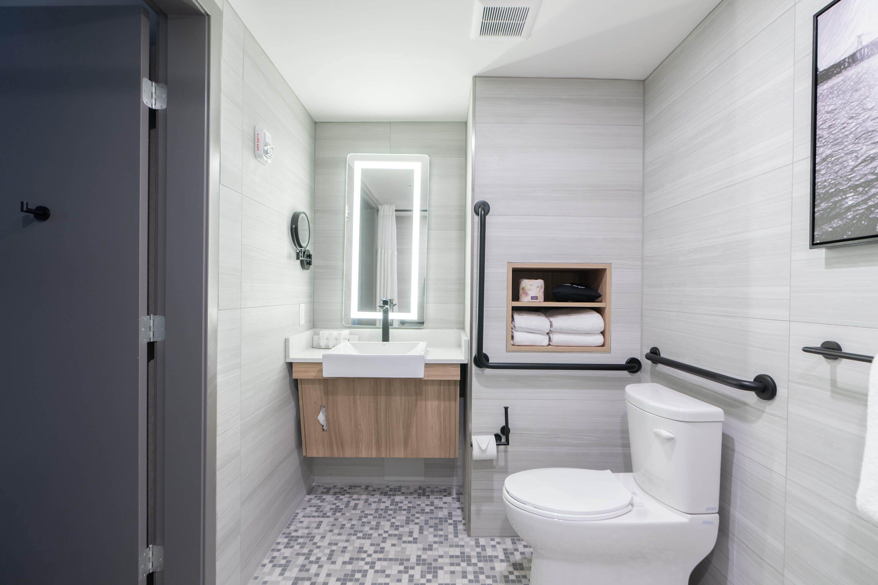 Un baño moderno con un lavamanos, espejo e inodoro blancos. Cuenta con una zona de ducha con puerta de vidrio y un perchero sobre el inodoro.