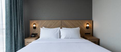 A modern hotel room with a large bed, two bedside tables with lamps, a wooden headboard, and a view of the city.