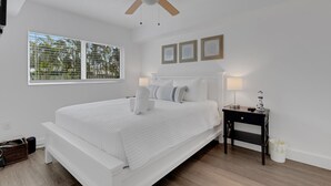 A modern bedroom with a large bed, a ceiling fan, a nightstand with a lamp, and a window with blinds. - Charming Beach Condo Steps to Beach | Heated Pool | Sleeps 4 (Bradenton Beach)