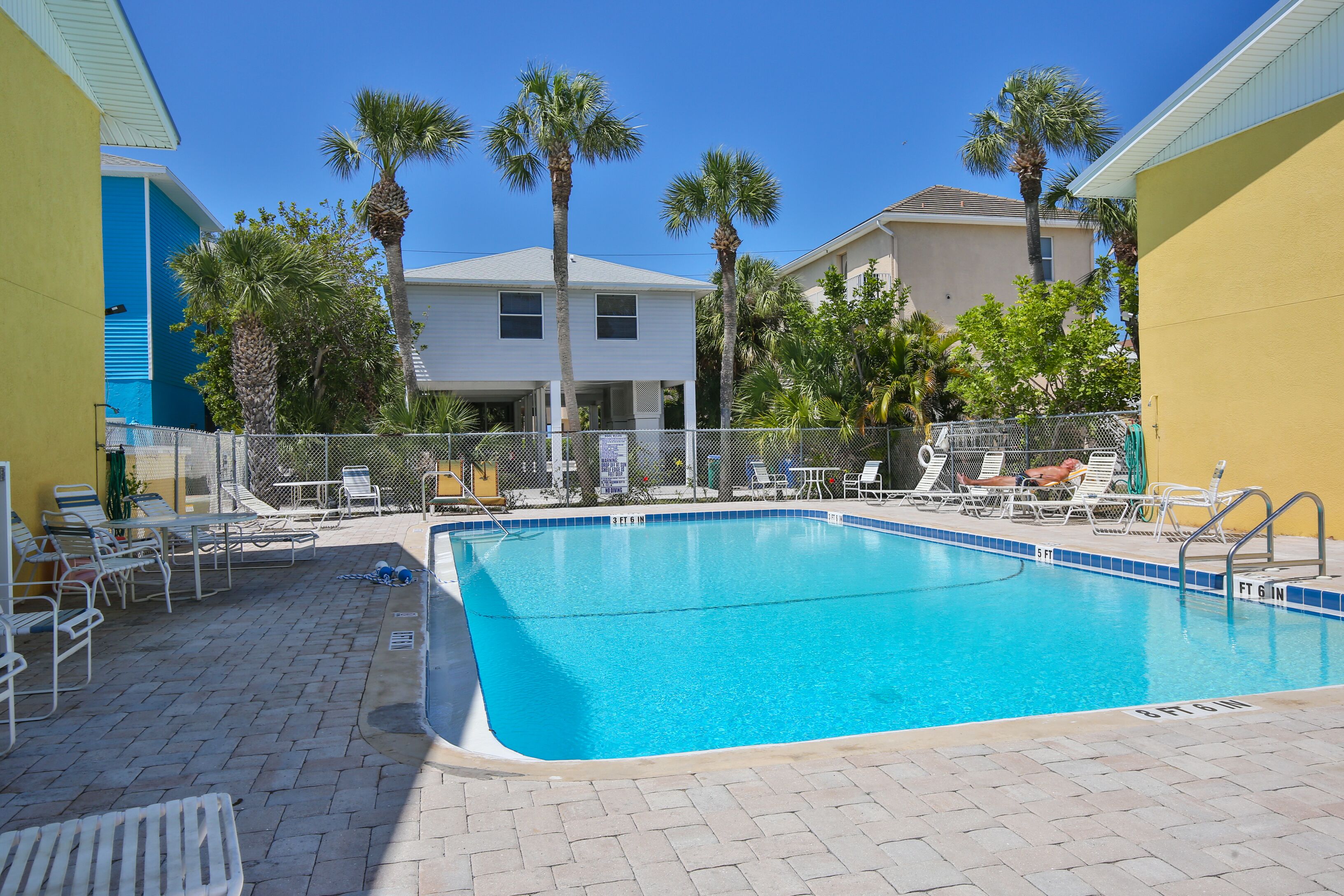 A swimming pool with lounge chairs and a fenced area, surrounded by palm trees and buildings.