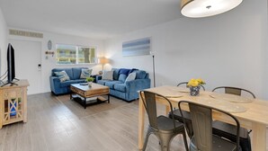 A modern living room with a blue sofa, a wooden coffee table, and a dining table with chairs. - Charming Beach Condo Steps to Beach | Heated Pool | Sleeps 4 (Bradenton Beach)