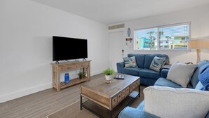A living room with a flat-screen TV, a wooden coffee table, a blue sofa, and a window with blinds. - Charming Beach Condo Steps to Beach | Heated Pool | Sleeps 4 (Bradenton Beach)