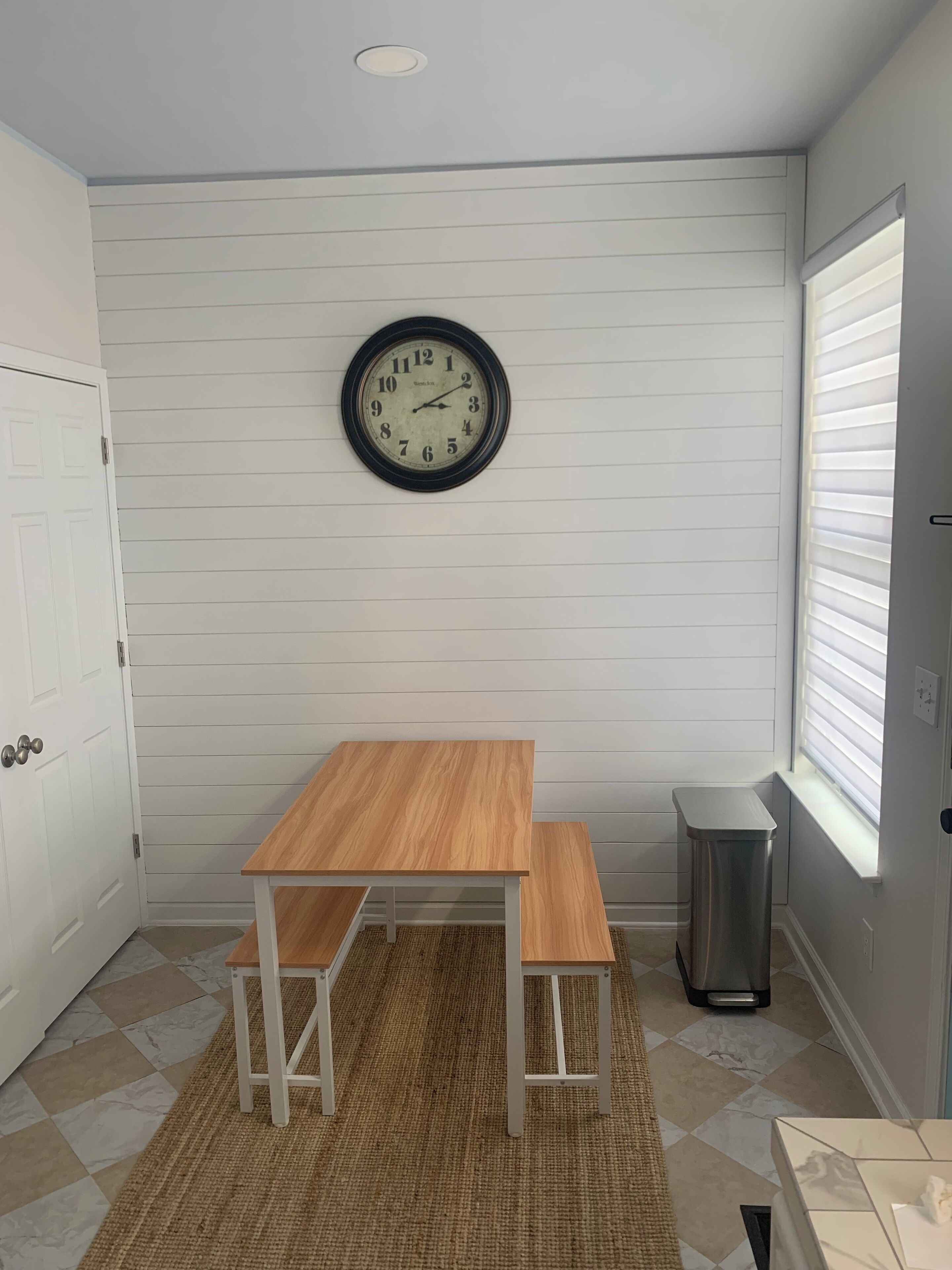 Una pequeña cocina con una mesa y banco de madera, un reloj en la pared y una ventana con persianas.