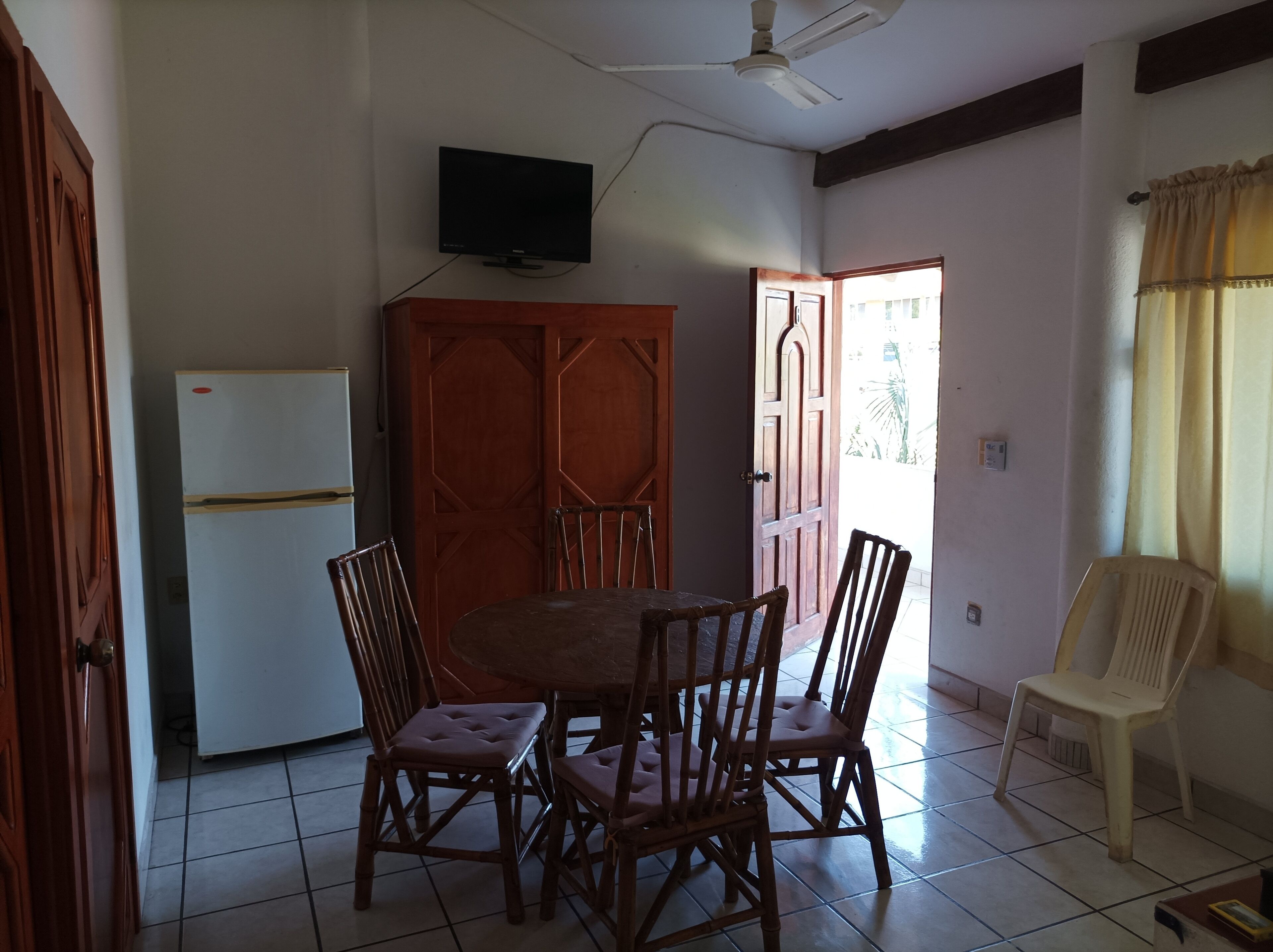Un coin repas avec une table ronde et quatre chaises, un ventilateur de plafond, un téléviseur fixé au mur et une porte donnant sur l’extérieur.