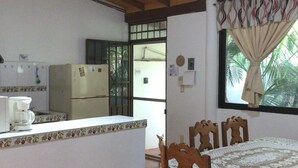 Une cuisine avec un coin repas, des poutres apparentes en bois au plafond, un réfrigérateur, une machine à café et une fenêtre avec des rideaux.