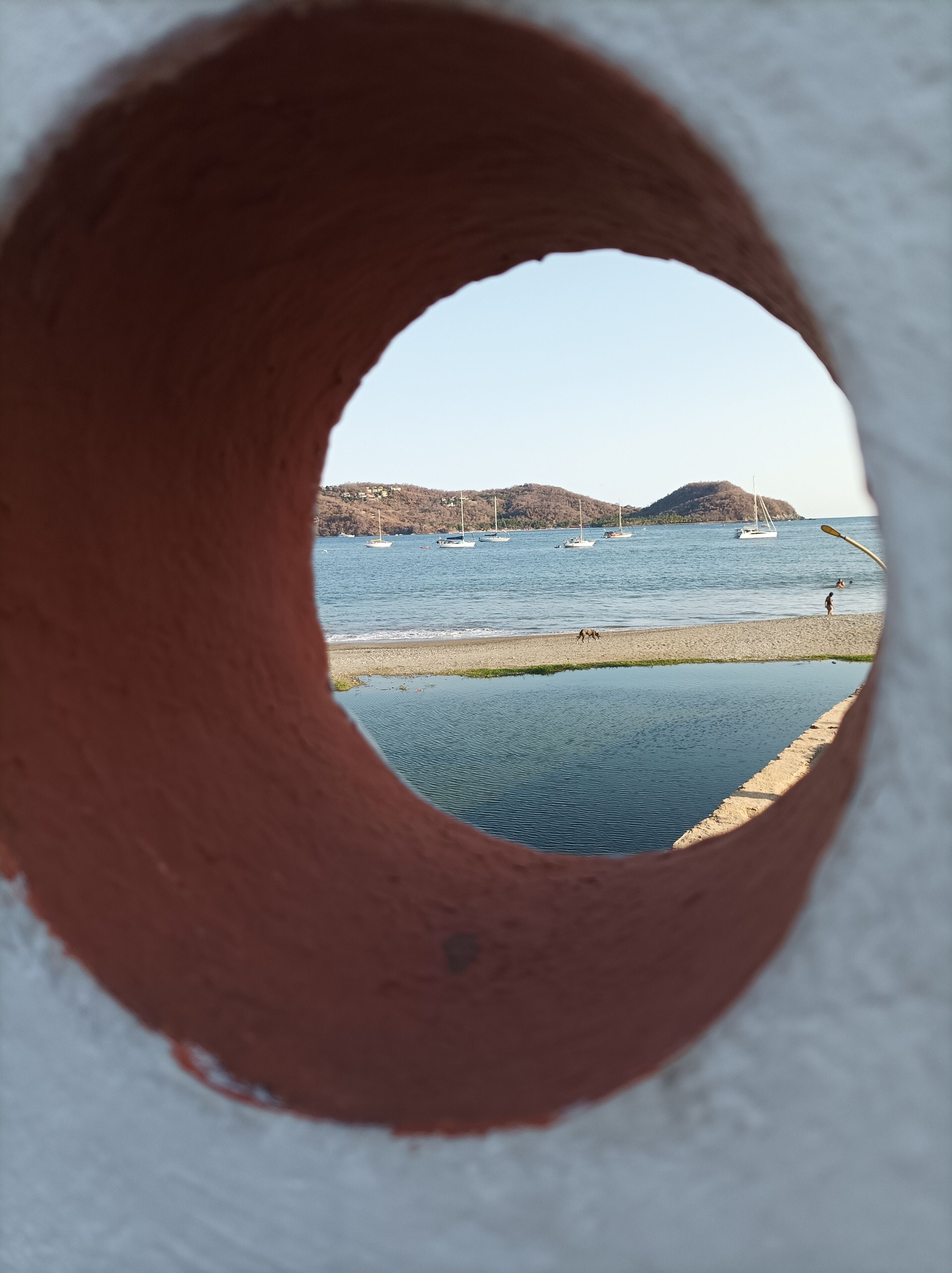 Uitzicht door een ronde opening, op een strand met boten en een heuvel in de verte.