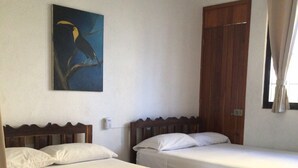 Une chambre d’hôtel avec deux lits simples, une tête de lit en bois, un tableau représentant un oiseau, une porte en bois et une fenêtre avec des rideaux.