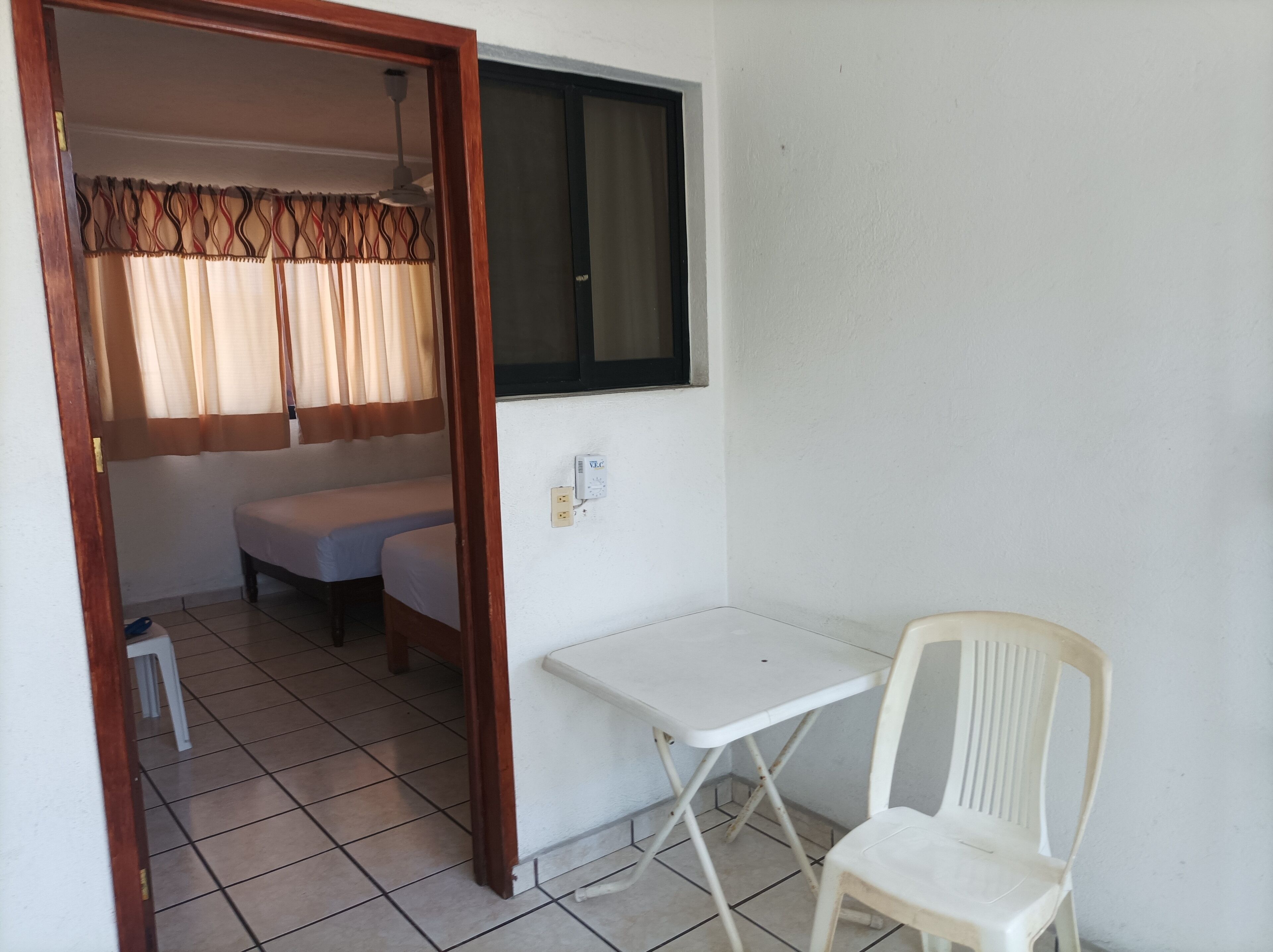 A small, clean room with a bed, a table, and a chair. There is a window with curtains, a power outlet, and a tiled floor.