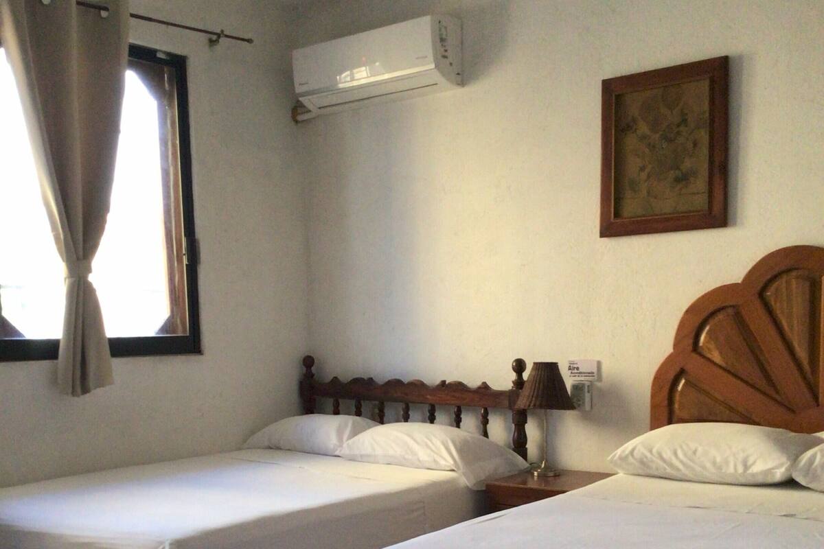 Une chambre d’hôtel avec deux lits simples, un ventilateur de plafond, un appareil de climatisation, une fenêtre avec des rideaux et un tableau encadré au mur.