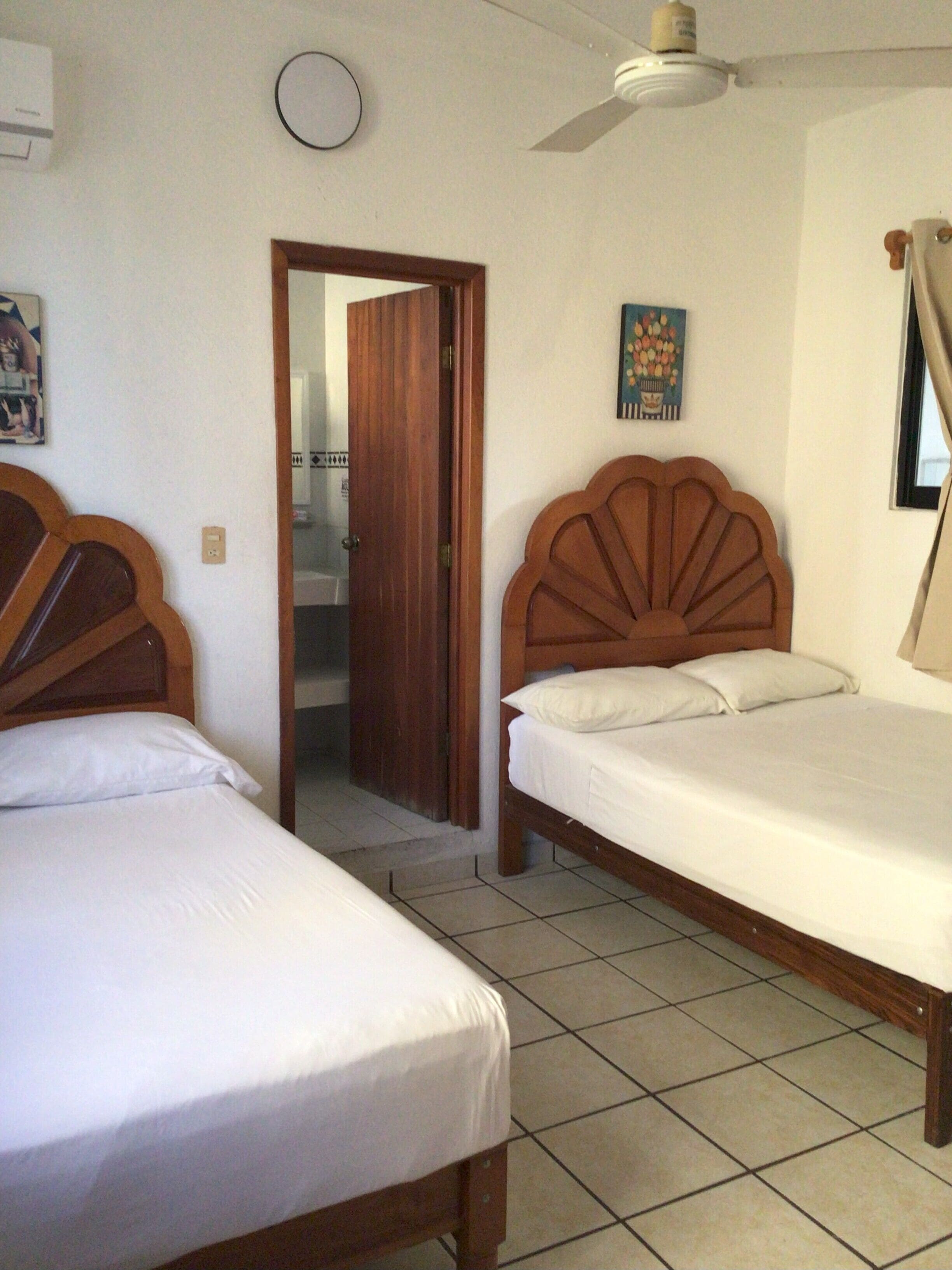 Una habitación con dos camas de madera, un ventilador de techo, una puerta que da a otra habitación y un cuadro colgado en la pared.