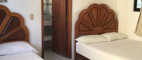 Una habitación con dos camas de madera, un ventilador de techo, una puerta que da a otra habitación y un cuadro colgado en la pared.