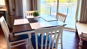 Un coin repas avec une table en bois, des chaises blanches, une plante en pot et une vue sur un lac, visible à travers de grandes fenêtres.