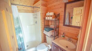 Une salle de bain rustique, aménagée dans une cabane en bois, comprenant des toilettes, une douche, un lavabo et une étagère avec des serviettes.