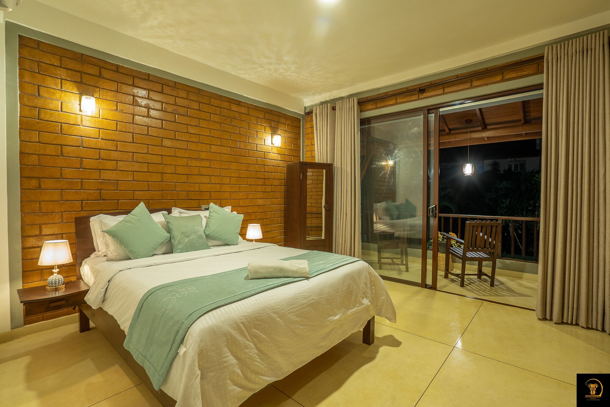 A hotel room with a large bed, a nightstand with a lamp, a wooden headboard, and a balcony with a chair.