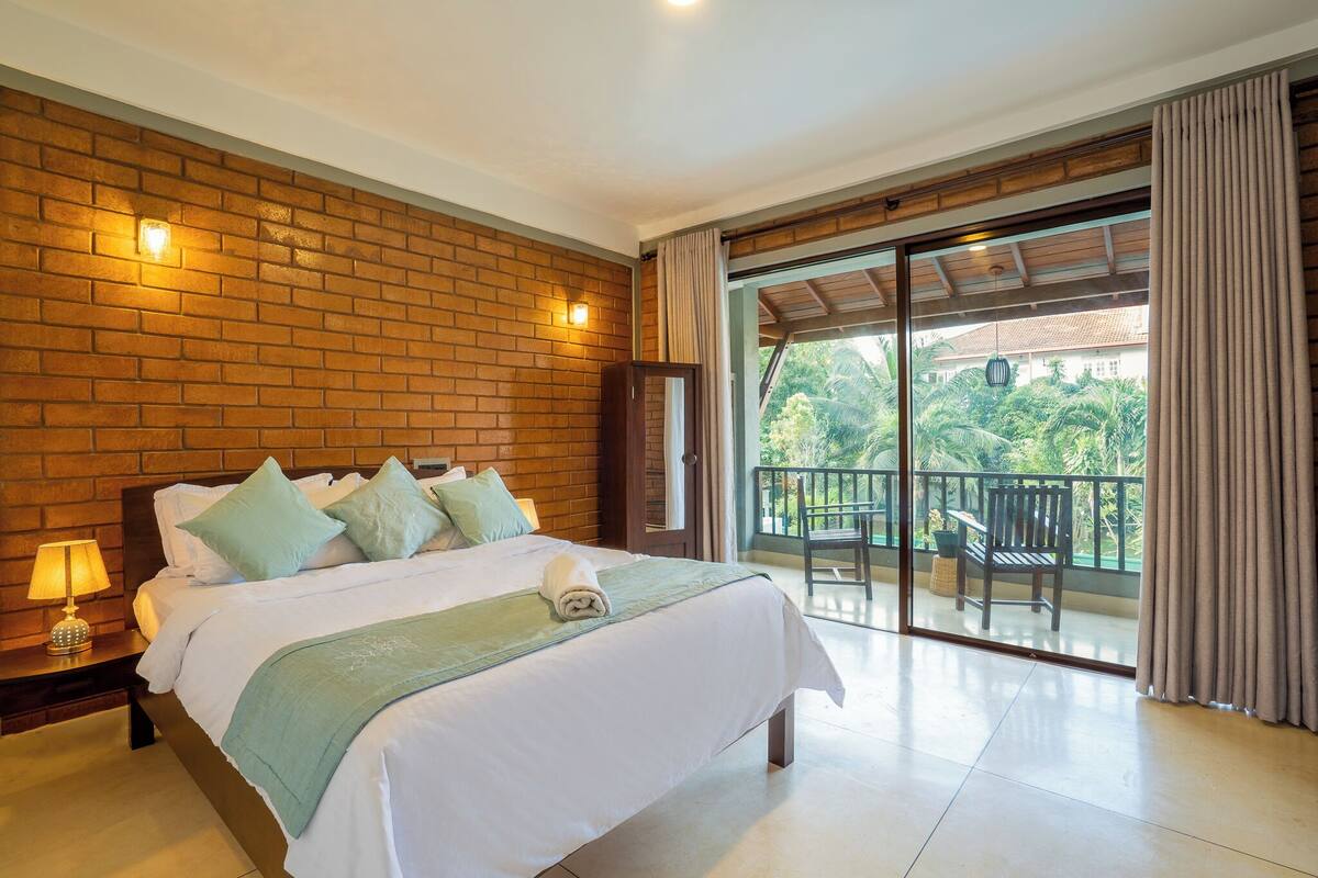 Quarto de hotel com parede de tijolos, uma cama grande com almofadas, mesa de cabeceira com abajur e varanda com cadeira e mesa.