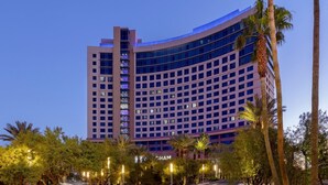 A high-rise hotel with a curved facade, multiple balconies, and a well-lit entrance area with palm trees.