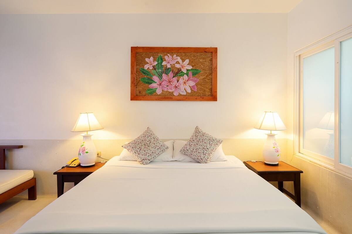 A hotel room with a large bed, two bedside lamps, a floral painting on the wall, and a window on the right.