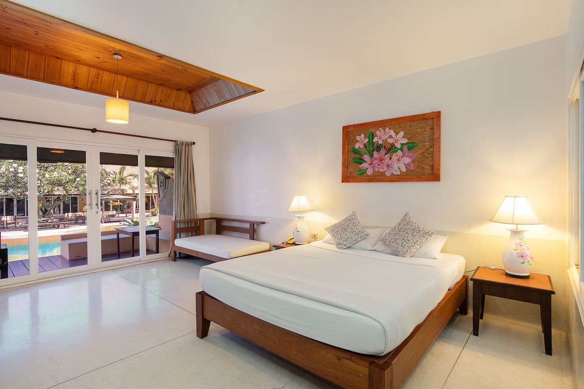 A bedroom with a large bed, two bedside tables with lamps, a painting on the wall, and a view of an outdoor pool area.