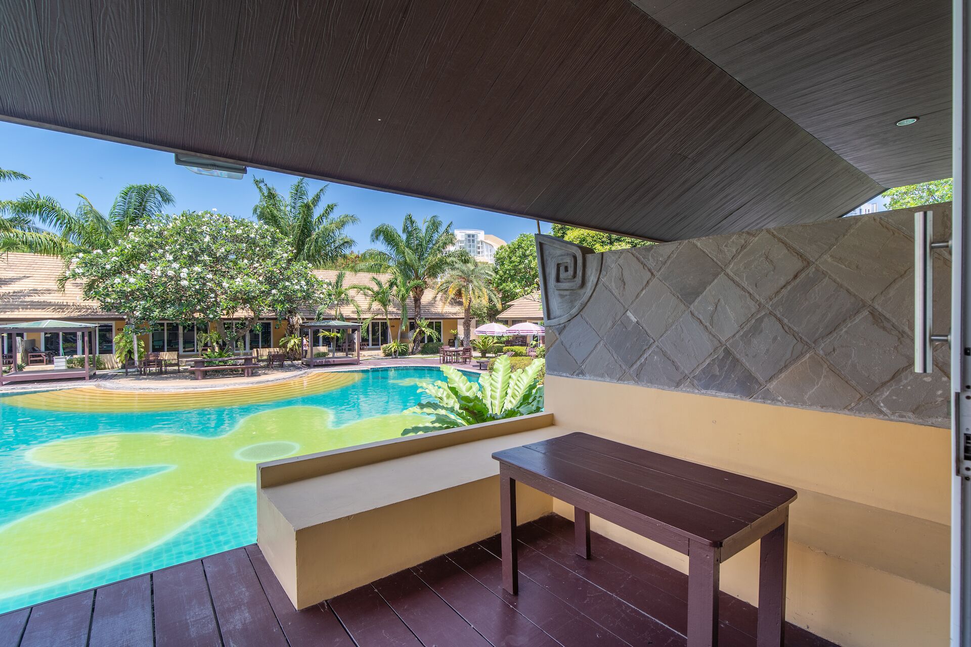 Zona junto a la piscina con terraza de madera, un banco y vistas a una piscina con palmeras y edificios de un complejo turístico al fondo.