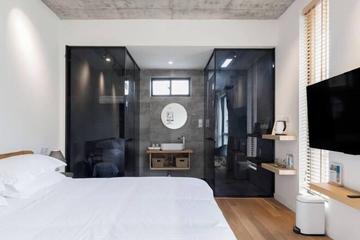 Una habitación de hotel moderna con cama, televisor, baño con espejo y lavamanos, y una ventana.