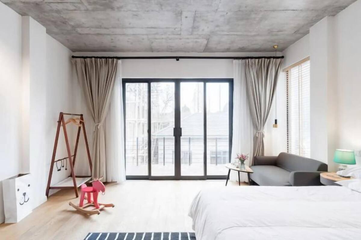 Un hotel moderno con una cama grande, un sofá gris, una mesita con una lámpara, un sillón mecedora y vistas a través de puertas de cristal.